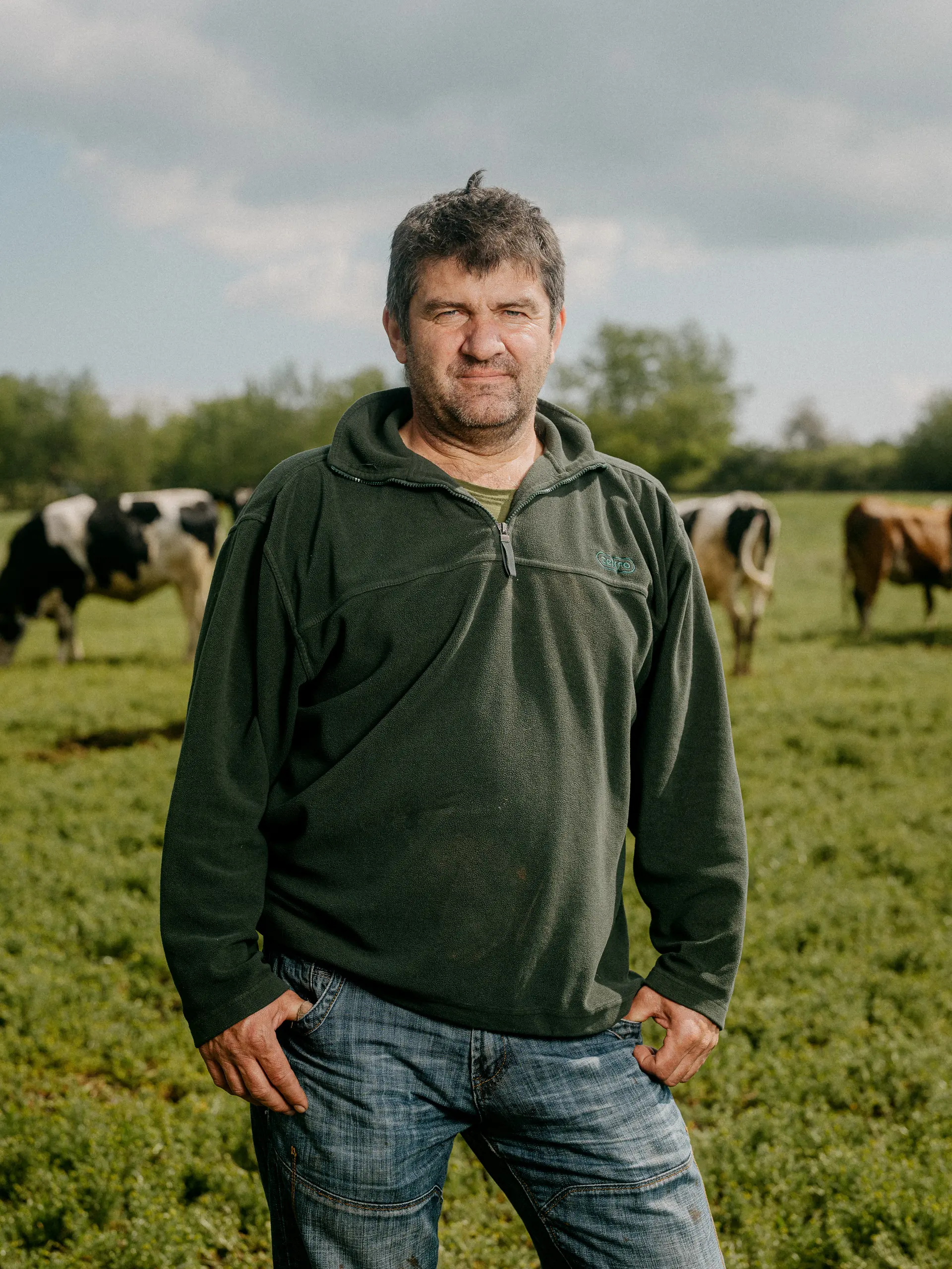 Farmář stojící na pastvině, krávy v pozadí.